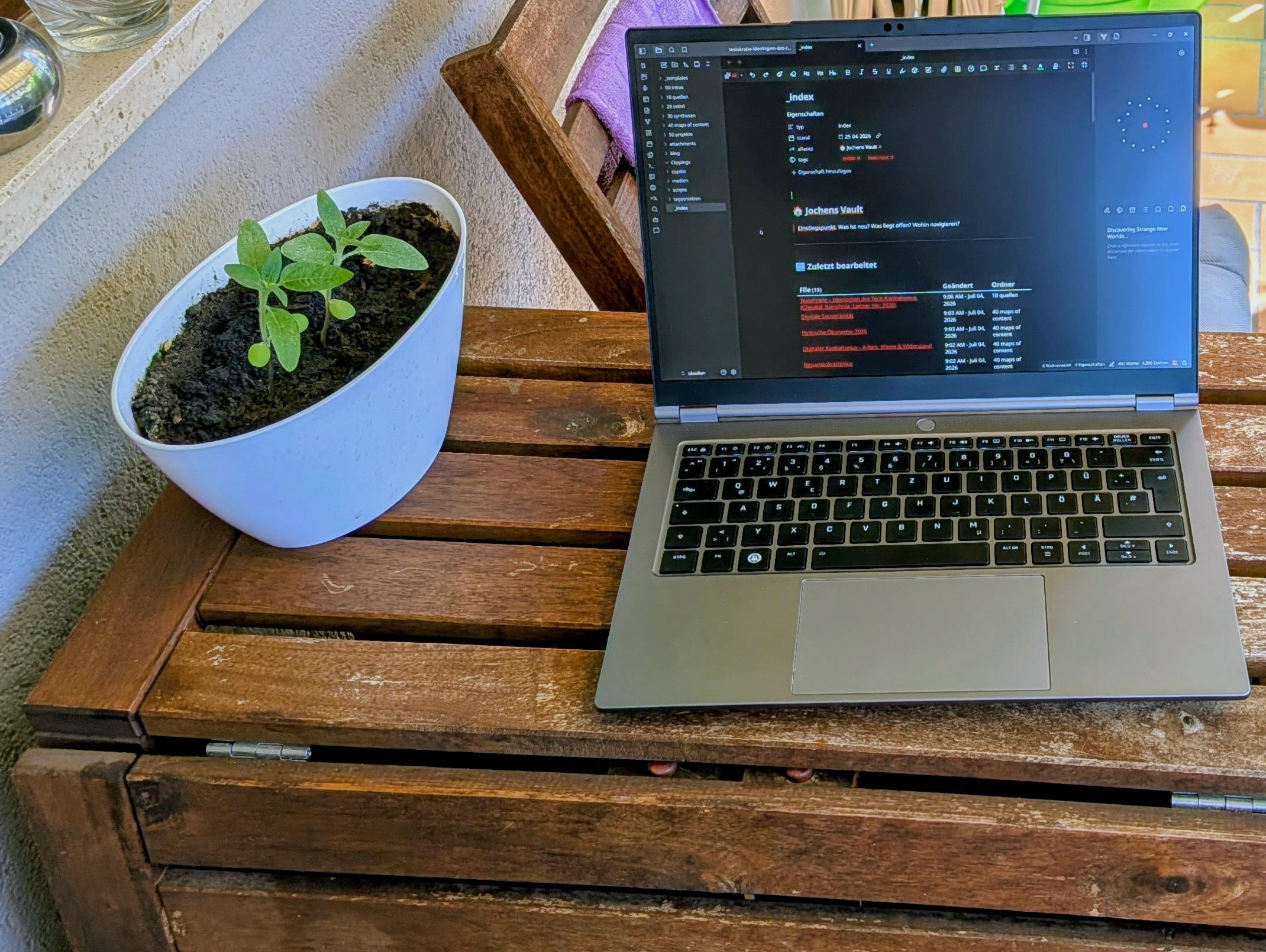 Aufgeklapptes Tuxedo InfinityBook Pro 14 auf einem Holztisch auf dem Balkon, auf dem Display der offene Obsidian-Vault, daneben ein Setzling im Topf